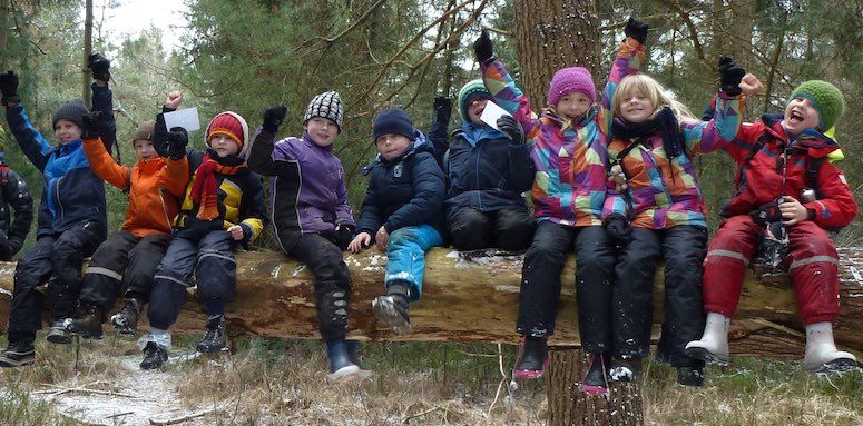 9 Kinder, die sich freuen und teilweise jubeln. Sie sitzen auf einem waaagerechten Baumstamm.