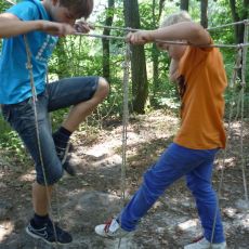 2 Kinder stehen kurz über der Erde in einem Parcours aus Seilen, die hängen und versuchen an einander vorbei zu klettern.