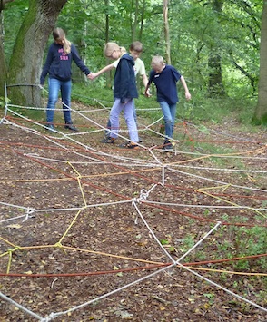 4 Kinder versuchen ein Spinnennetz aus Seilen, das ungefähr 30 cm über dem Boden angebracht ist, zu überwinden.