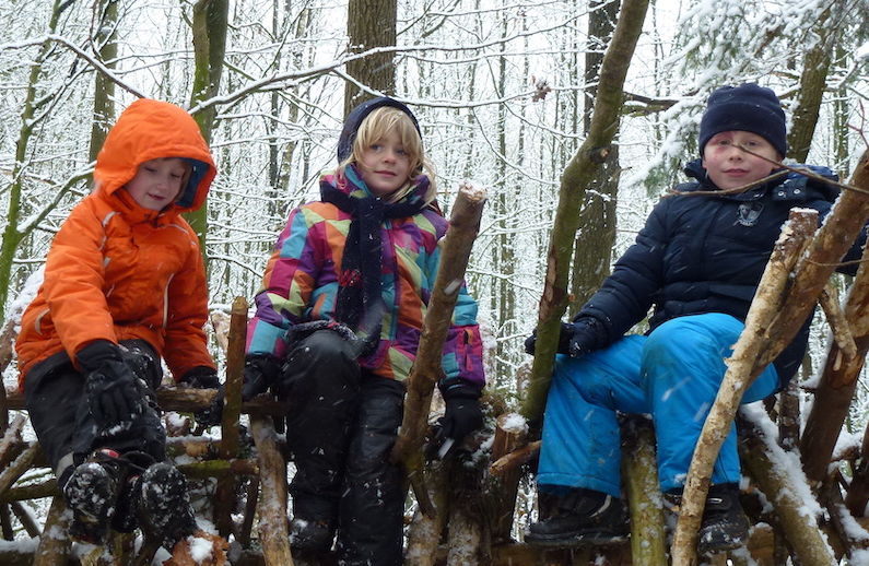 3 Kinder sitzen auf einen selbstgeebauten Unterstand aus Ästen. Im Hintergrund sind Bäume. Es liegt Schnee. 